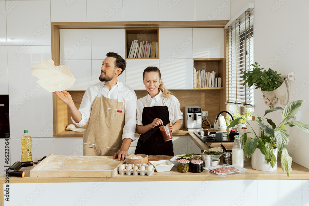 Happy man rolling homemade dough with hands throwing up baking cooking homemade pizza in modern light kitchen. Beautiful blonde woman with ponytail mixing ketchup in measuring cup. Happy man rolling homemade dough with hands throwing up baking cooking homemade pizza in modern light kitchen. Beautiful blonde woman with ponytail mixing ketchup in measuring cup.