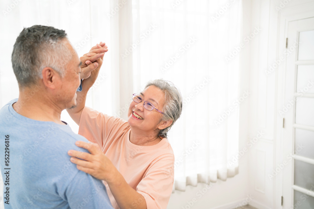 Obraz premium Romantic and cheerful Asian well-being senior couple enjoy dancing and holding hands to music together with smiles and happiness in the living room. Senior retirement activity at home.