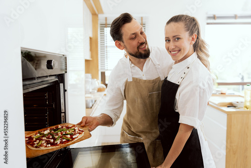 Lovely adorable couple in modern light kitchen taking fresh kneaded pizza in oven. Having fun together. Handsome husband lovely loking at beautiful wife.