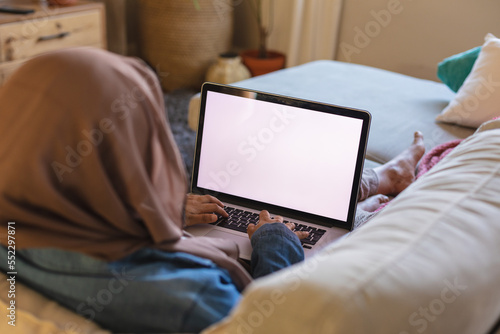 Biracial woman wearing hijab, laying on sofa in living room and using laptop with copy space