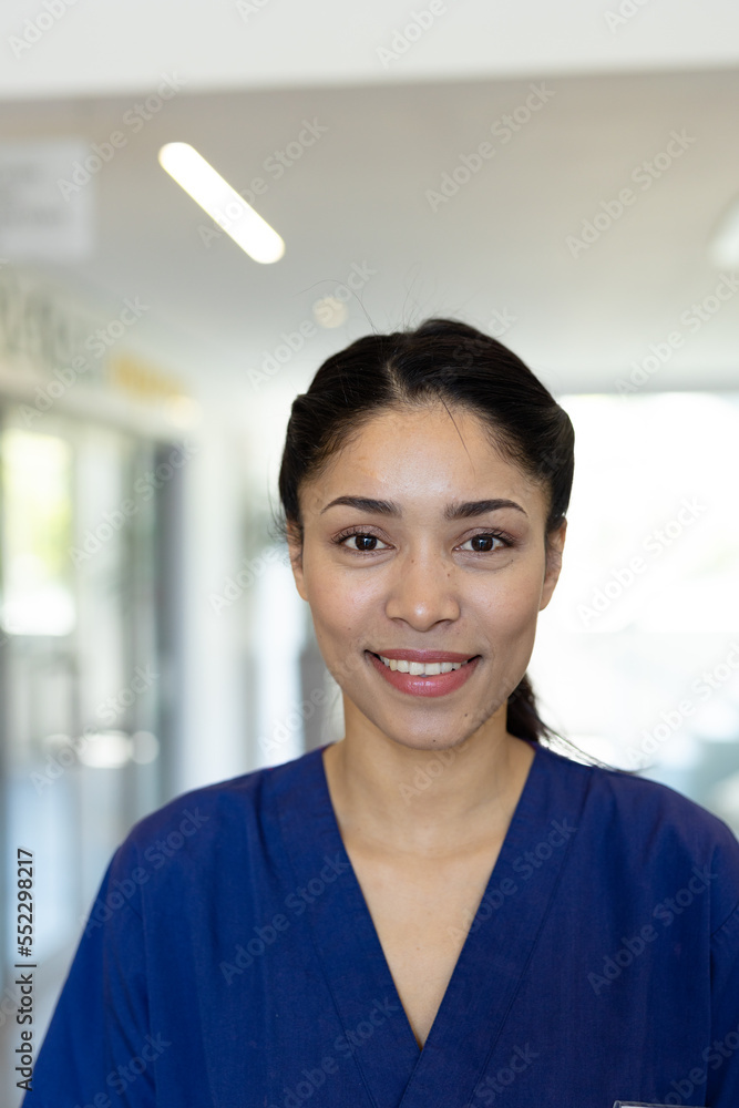 Vertical portrait of smiling biracial female healthcare worker in hospital corridor, with copy space