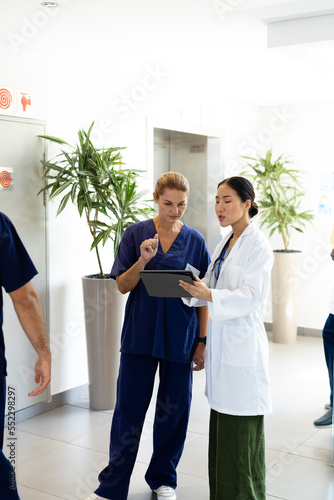 Vertical of two diverse female doctors with tablet talking in hospital corridor, with copy space