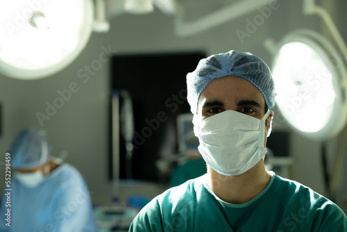 Portrait of biracial male surgeon in surgical cap and mask in operating theatre with copy space