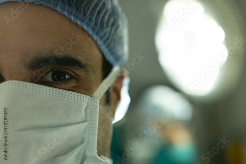 Portrait of biracial male surgeon in surgical cap and mask in operating theatre with copy space