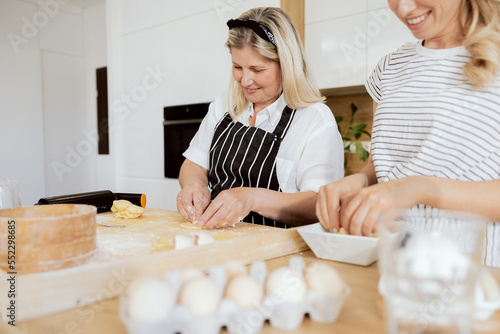 Elderly woman kneading dough with adorable daughter filling in dough with potato.Woman preparing cooking homemade varennyky on wooden surface. Senior mother enjoying momet with young lady. photo