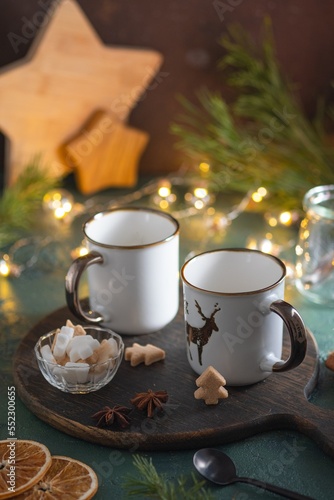 Two empty white mugs on a wooden board on a green concrete background in a Christmas composition. Prepared serving for hot drinks.