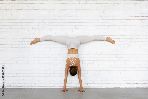 Young woman trainer practicing yoga doing Adho Mukha Vrikshasana exercise with Samakonasana, handstand with transverse splits with legs against the wall, inverted asana, exercising in white clothes photo