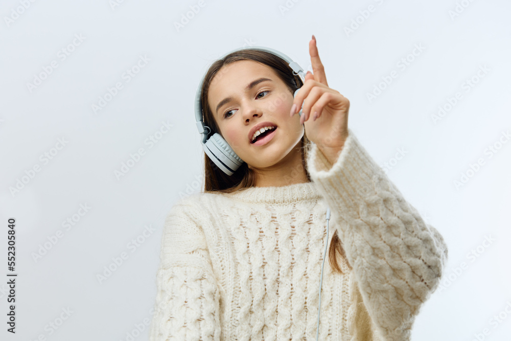 a happy, joyful woman stands on a light gray background in a stylish sweater and happily dances pointing her index finger up while listening to music in headphones a happy, joyful woman stands on a light gray background in a stylish sweater and happily dances pointing her index finger up while listening to music in headphones