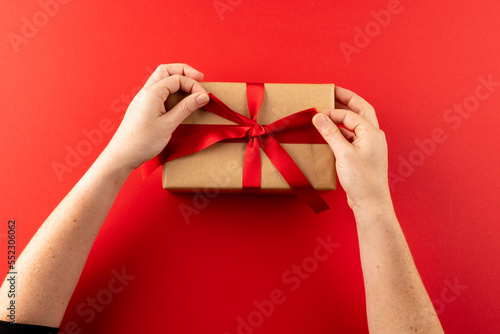Overhead of hands tying gift in brown paper with red ribbon, on red background with copy space © vectorfusionart