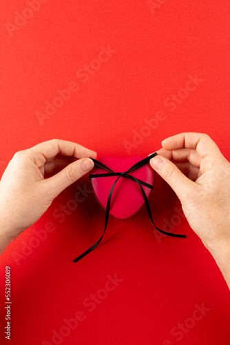 Vertical of hands tying red heart gift box with black ribbon  on red background with copy space