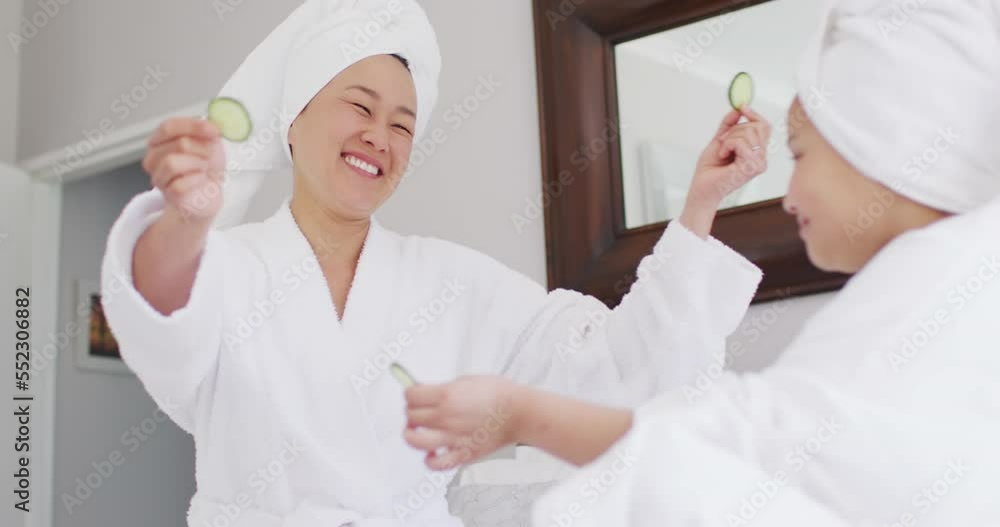 Video of happy asian mother and daughter in robes doing cucumber mask and having fun Video of happy asian mother and daughter in robes doing cucumber mask and having fun