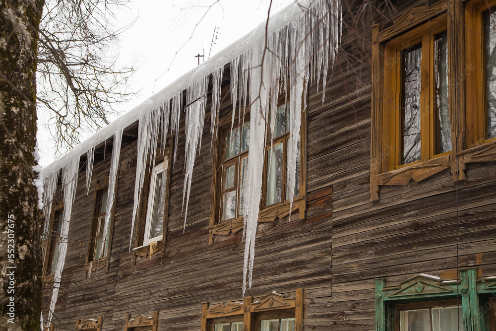 Long, melting icicles hang on the edge of the roof, winter or spring. Log wall of an old wooden house with windows. Large cascades of icicles in smooth, beautiful rows. Cloudy winter day, soft light.