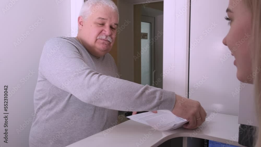 Elderly man client signing paper at reception desk In hospital. Man fills out a questionnaire. Patient being attended by a nurse in a medical consultation. Patient and receptionist in dental clinic Elderly man client signing paper at reception desk In hospital. Man fills out a questionnaire. Patient being attended by a nurse in a medical consultation. Patient and receptionist in dental clinic