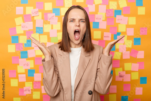 Quadro em tela Photo of despair angry woman wearing beige jacket standing against yellow wall covered with stickers, standing with raised arms, screaming with aggression, arguing with somebody