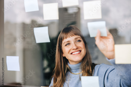 Cheerful young businesswoman sticking adhesive notes to a glass wall photo