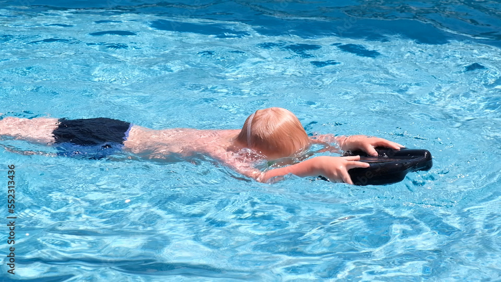 Caucasian child learning to swim in the pool in summer wearing swimming goggles and a swim board.