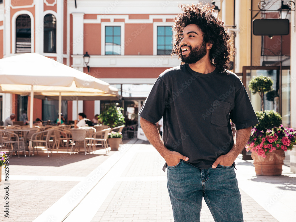 Fototapeta premium Handsome smiling hipster model.Sexy unshaven Arabian man dressed in summer clothes. Fashion male with long curly hairstyle posing in the street at sunset. Cheerful and happy