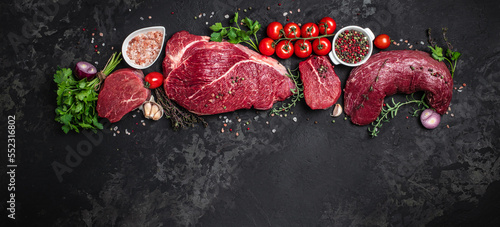 Assortment of raw cuts of raw beef meat steaks with spices on a dark background. Long banner format. top view