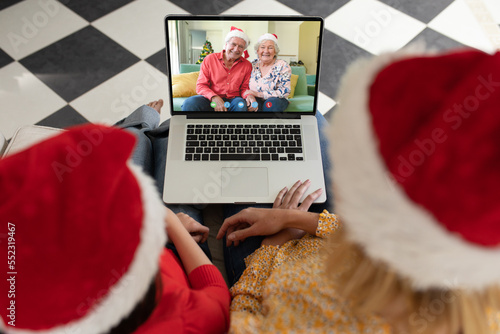 Diverse mother and daughter with santa hats having video call with happy senior caucasian couple