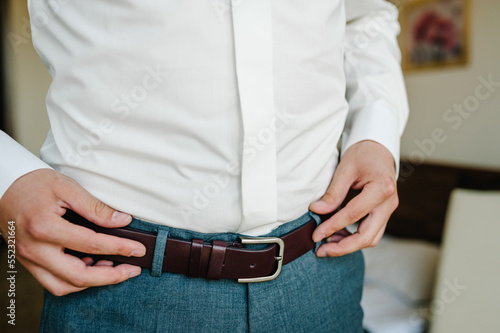 A man in pants and shirt buttoned a brown leather trouser belt. Groom Dress up a belt with buckle. Businessman wear leather stylish belt. New classic belt. Close up. © Serhii