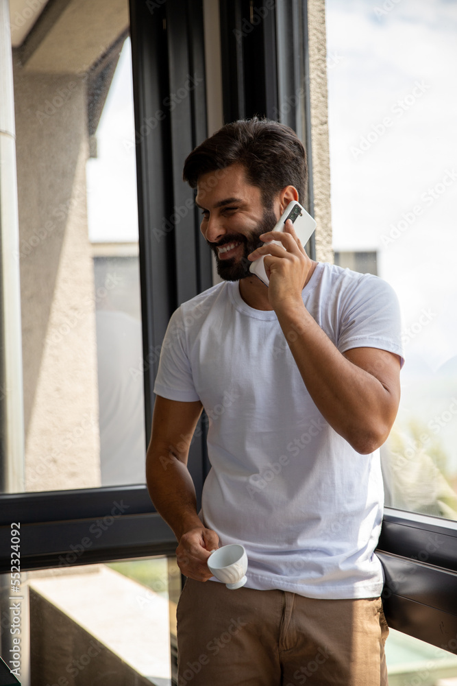 Fototapeta premium handsome man answering his phone and smiling, man holds cup of coffee and answers his smartphone smiling