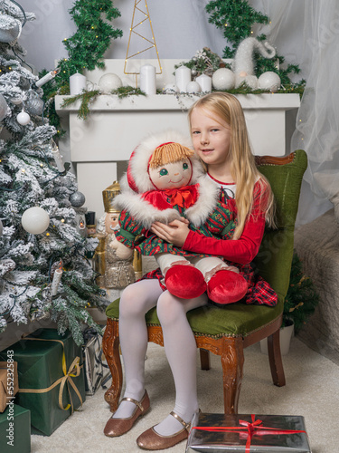 A little blonde girl is sitting on a chair in a Christmas interior. The girl is holding a doll in her hands. Magical Christmas time. Vertical photo.