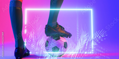 Low section of african american male player with leg on ball by illuminated rectangle and plants