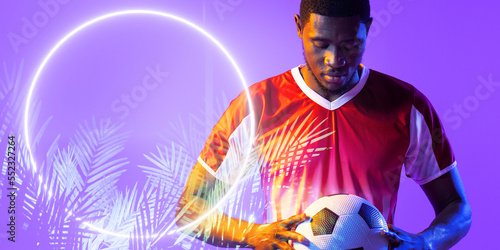 African american male soccer player holding soccer ball by illuminated circle and plants, copy space