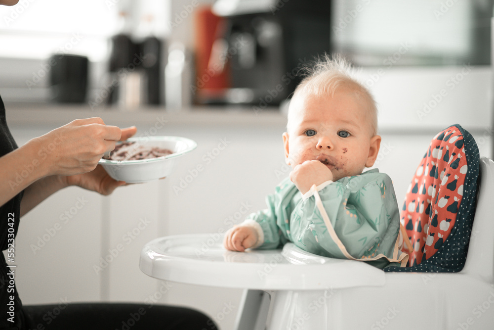 Mother spoon feeding her baby boy child in baby chair with fruit puree in kitchen at home. Baby solid food introduction concept Mother spoon feeding her baby boy child in baby chair with fruit puree in kitchen at home. Baby solid food introduction concept