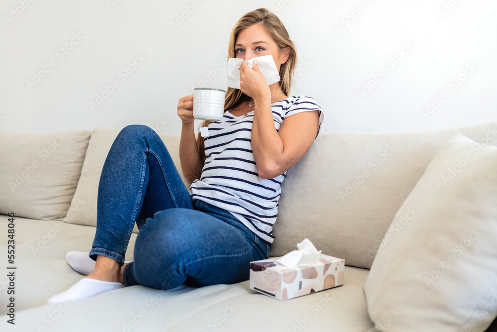 Sick young woman blowing running nose, feeling unhealthy and ill, upset female holding paper napkin, handkerchief, holding tea or coffee mug, sitting on couch at home. Sick young woman blowing running nose, feeling unhealthy and ill, upset female holding paper napkin, handkerchief, holding tea or coffee mug, sitting on couch at home.