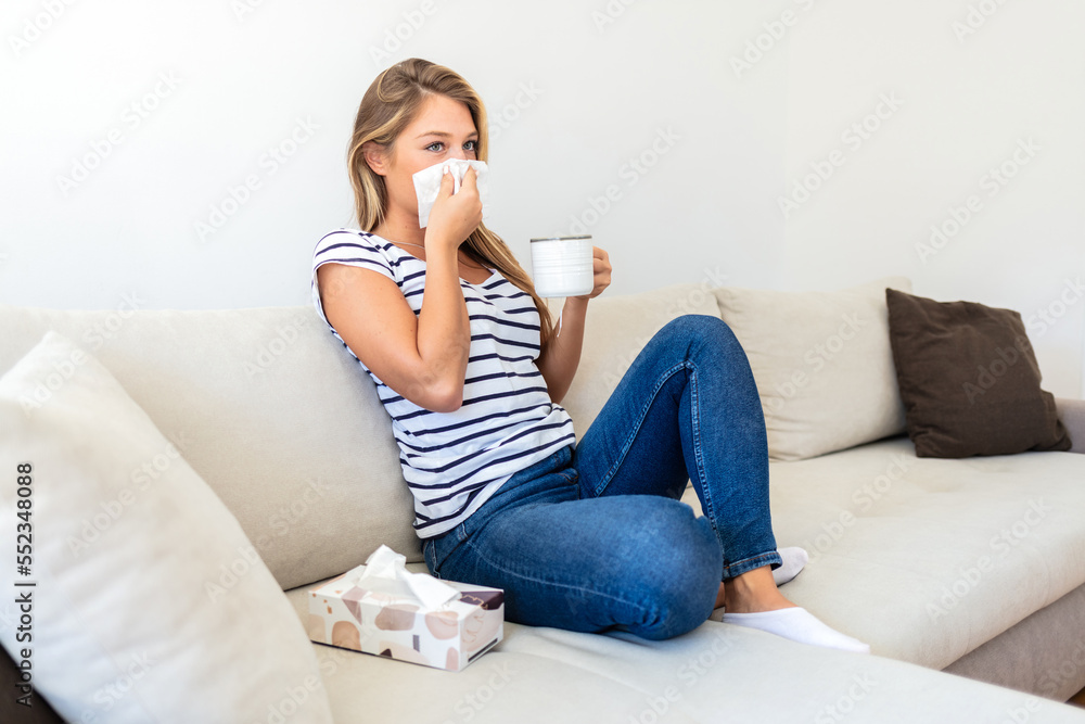 Young woman with cold blowing her runny nose. Unwell girl sneezing in a tissue in the living room. Sick young woman sneezing into a paper tissue while holding hot tea in domestic interior. Young woman with cold blowing her runny nose. Unwell girl sneezing in a tissue in the living room. Sick young woman sneezing into a paper tissue while holding hot tea in domestic interior.