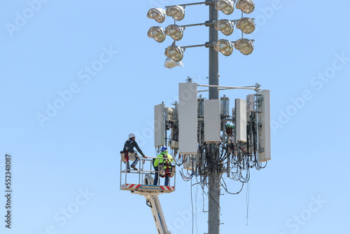 Telecom workers operate from a lift to service cellular antennas and cabling on a high light pole, illustrating mobile network infrastructure maintenance and high-elevation technical labor