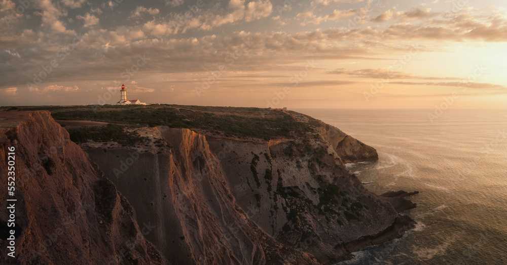 Warm summer cloudy sunset seascape with massive cliffs and sea on foreground, and a lit hexagonal eighteenth century lighthouse on background, Colour Photo, Cabo Espichel, Sesimbra, Setúbal, Portugal