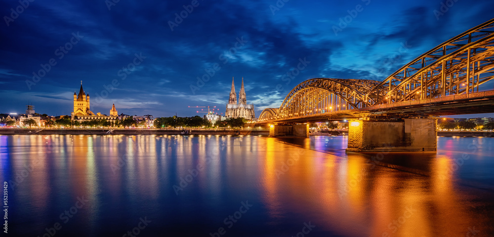 Fototapeta premium Panorama of illuminated bridge over Rhein river whith trains and tourists passing by and the Cologne Cathedral in the evening at the blue hour. Germany city life