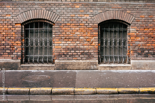 Frontal view of a red brick wall with two barred windows. In front of it a wet sidewalk and in front of it a puddle with reflection