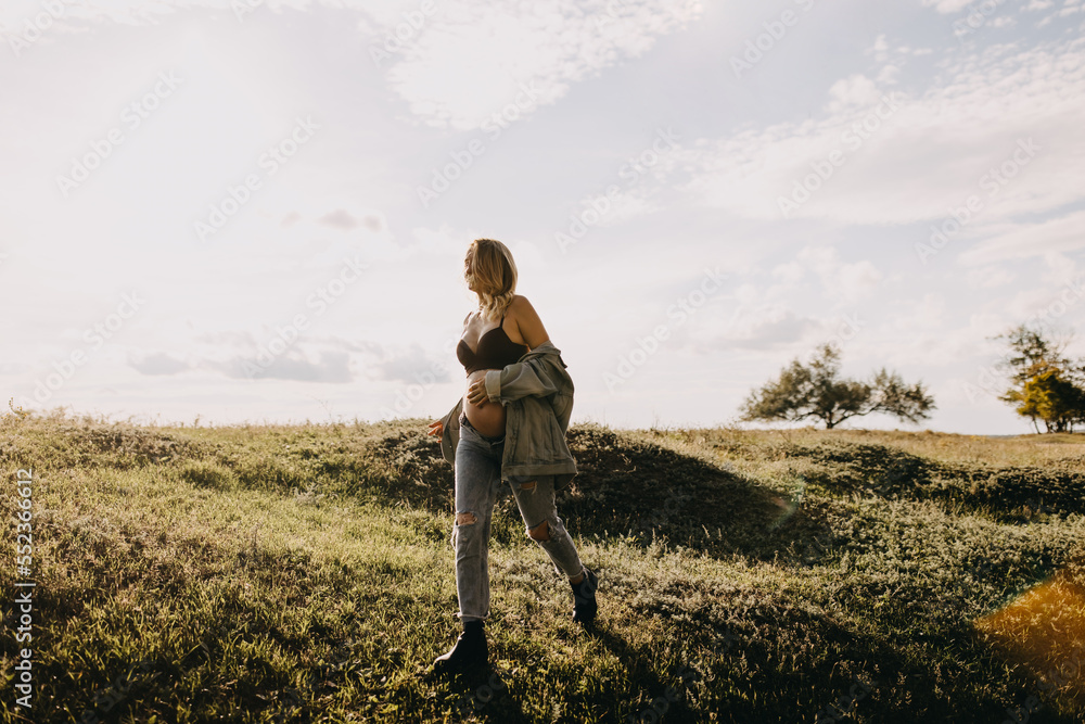 Blonde pregnant woman wearing a denim jacket and jeans, walking in an open field.