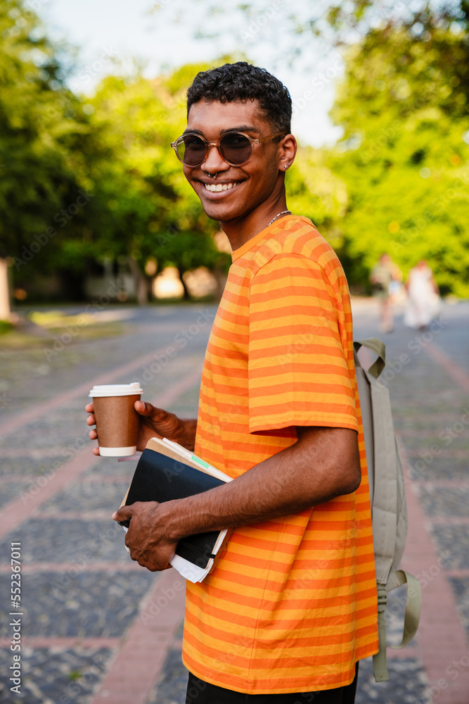 Obraz premium Young african man student holding coffee and books while standing outdoors