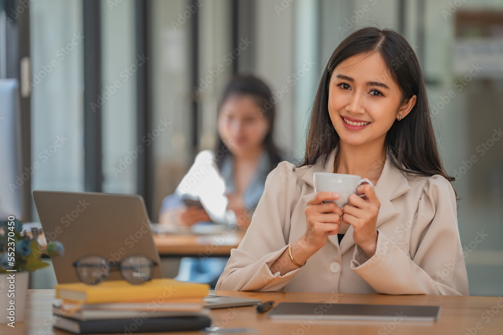 Beautiful woman holding a coffee cup and using a laptop while sitting at her working place in an office. Beautiful woman holding a coffee cup and using a laptop while sitting at her working place in an office.