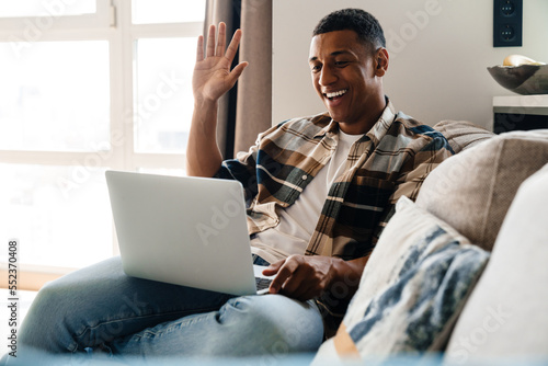 Young african man having video call via laptop and waving hand while sitting on couch