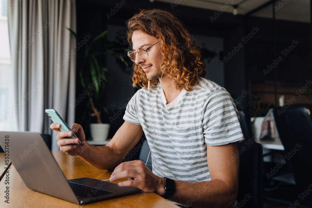Young manager using smartphone while working on laptop computer sitting at table Young manager using smartphone while working on laptop computer sitting at table