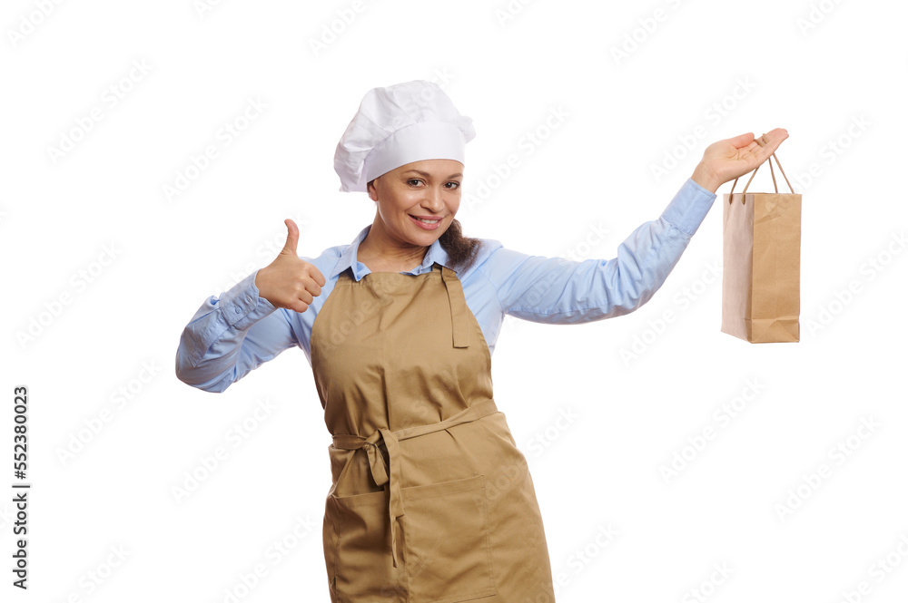 Pretty woman waitress or baker wearing chef hat and beige apron, holding a disposable paper bag with take out food and shows thumb up, cutely smiling looking at camera, isolated over white background Pretty woman waitress or baker wearing chef hat and beige apron, holding a disposable paper bag with take out food and shows thumb up, cutely smiling looking at camera, isolated over white background