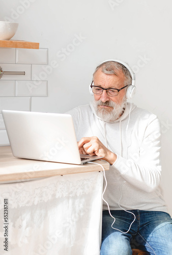 Portrait handsome old man dressed in hoodie and eyeglasses is using laptop and smiling while listen music sitting on couch at home
