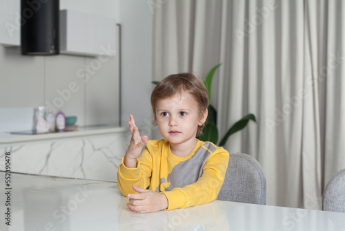 Portrait of adorable cute child boy in yellow sweater sitting by the table and raising his hand up on white studio background