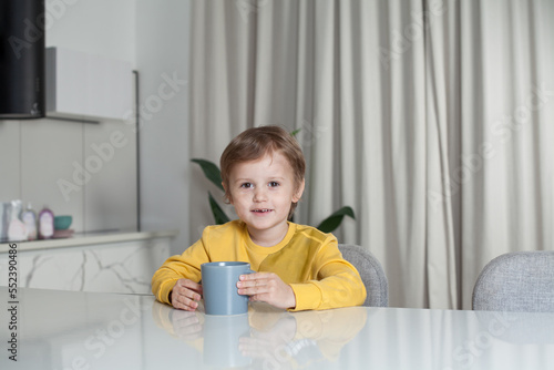 Happy cute child boy in yellow sweater sitting by the table and holding mug on white curtain background