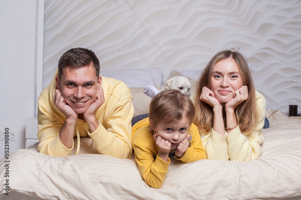 Portrait of lovely family, friendly parents, mom and dad with adorable child son and white dog laiyng on the bed