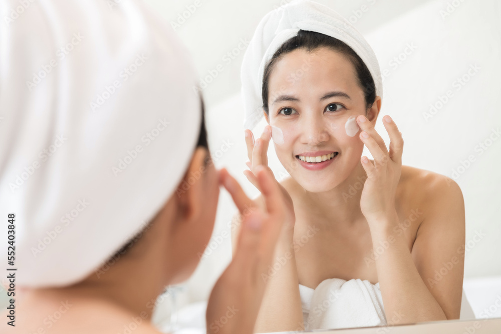 Happy young Asian woman applying face lotions while wearing a towel and touching her face in bathroom Happy young Asian woman applying face lotions while wearing a towel and touching her face in bathroom
