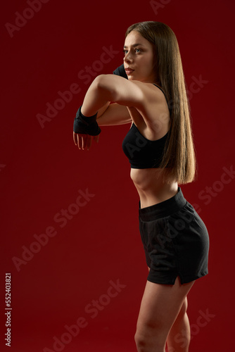 Beautiful female trainer showing kick exercise on red background. Seductive brunette woman training in black sport bandages in studio. Concept of martial arts. © serhiibobyk