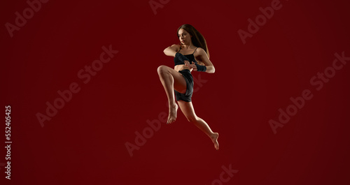 Side view of brunette athletic woman with long hair jumping high  on red studio background. Pretty strong female training in air. Concept of martial arts.