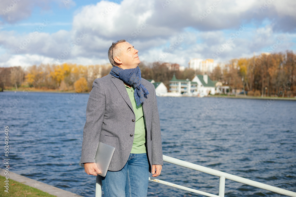 Portrait of happy middle-aged man standing with closed eyes on bank of river in city, enjoying sunlight, holding laptop.
