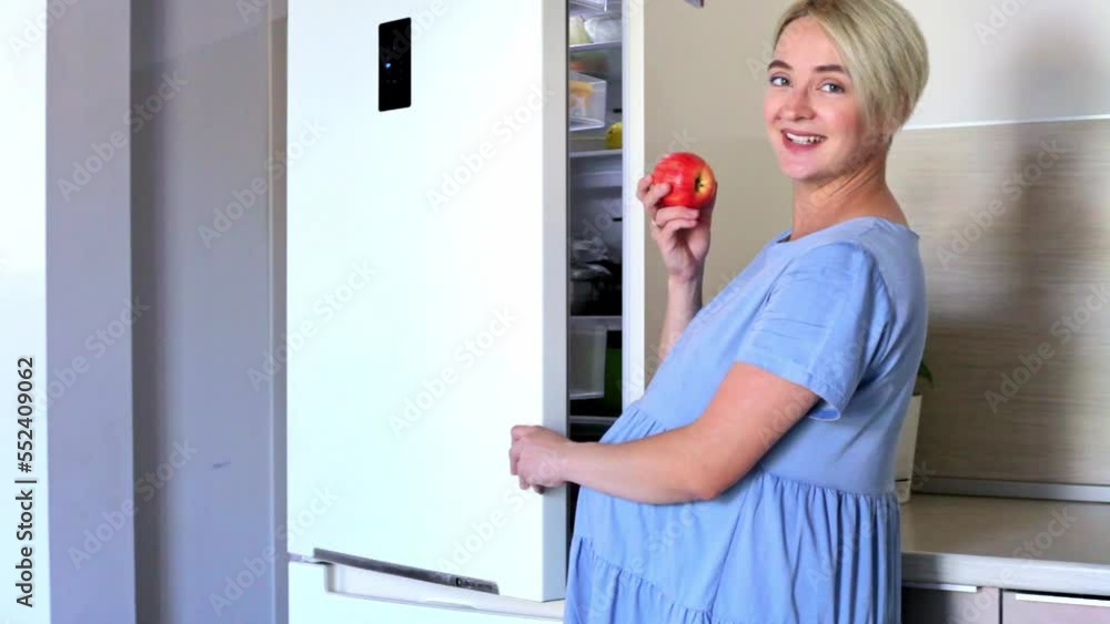 a young pregnant woman in a blue dress with a short haircut opens the refrigerator and leaves a red apple a young pregnant woman in a blue dress with a short haircut opens the refrigerator and leaves a red apple